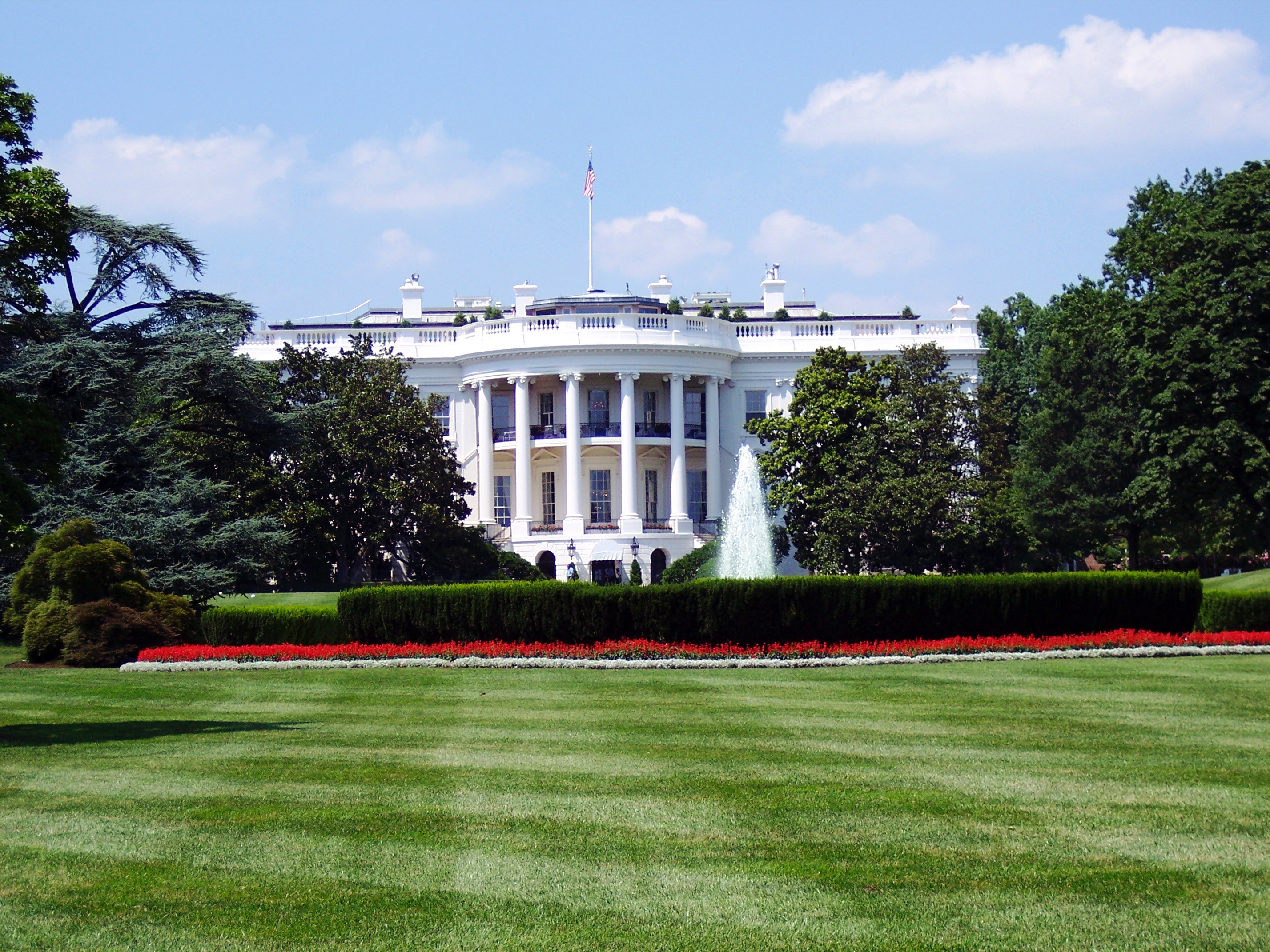 Exterior of the White House and it's lawn on a sunny day with a blue sky.