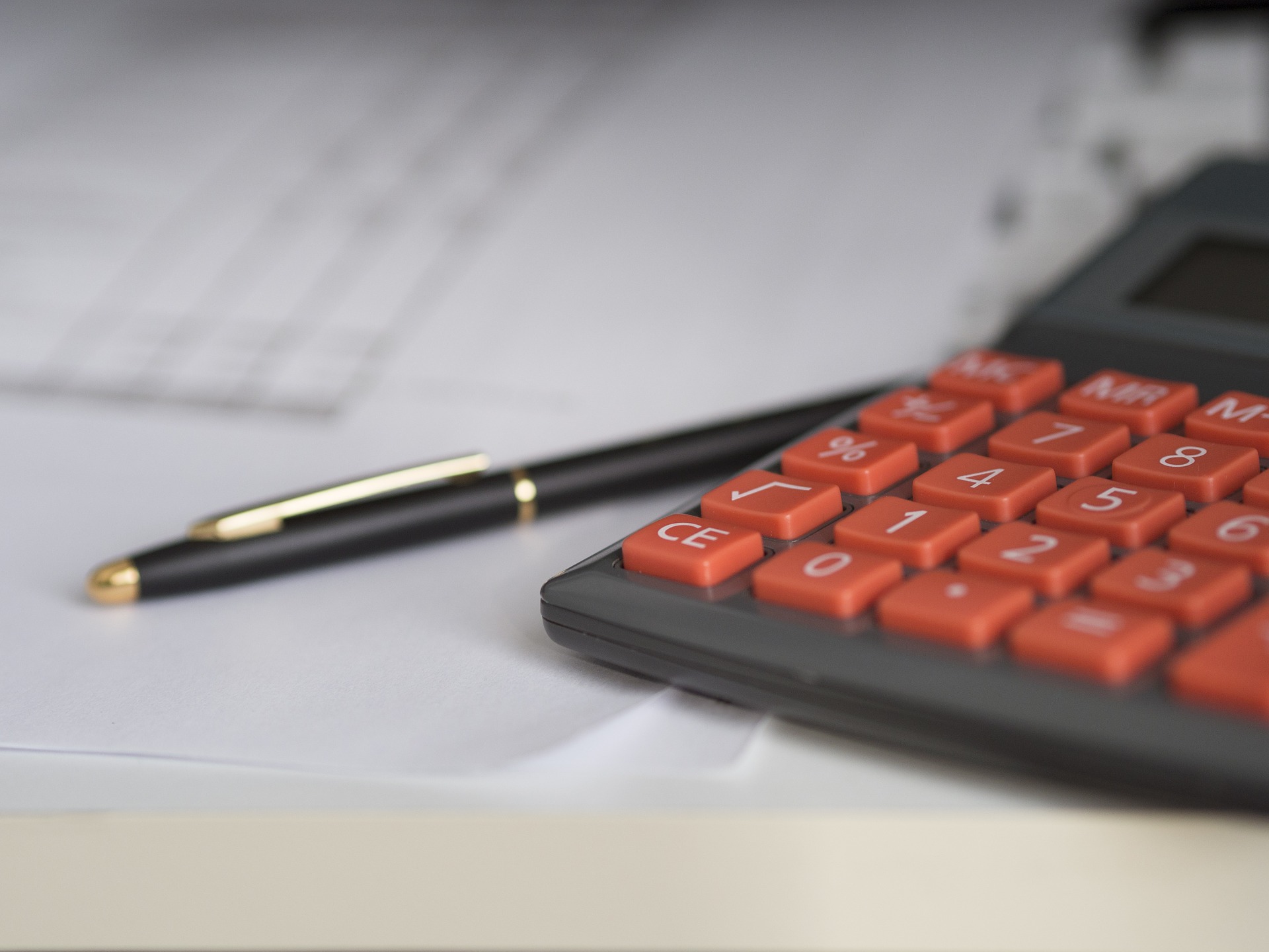 Black calculator with red buttons lays next to pen, on top of a pile of white papers.