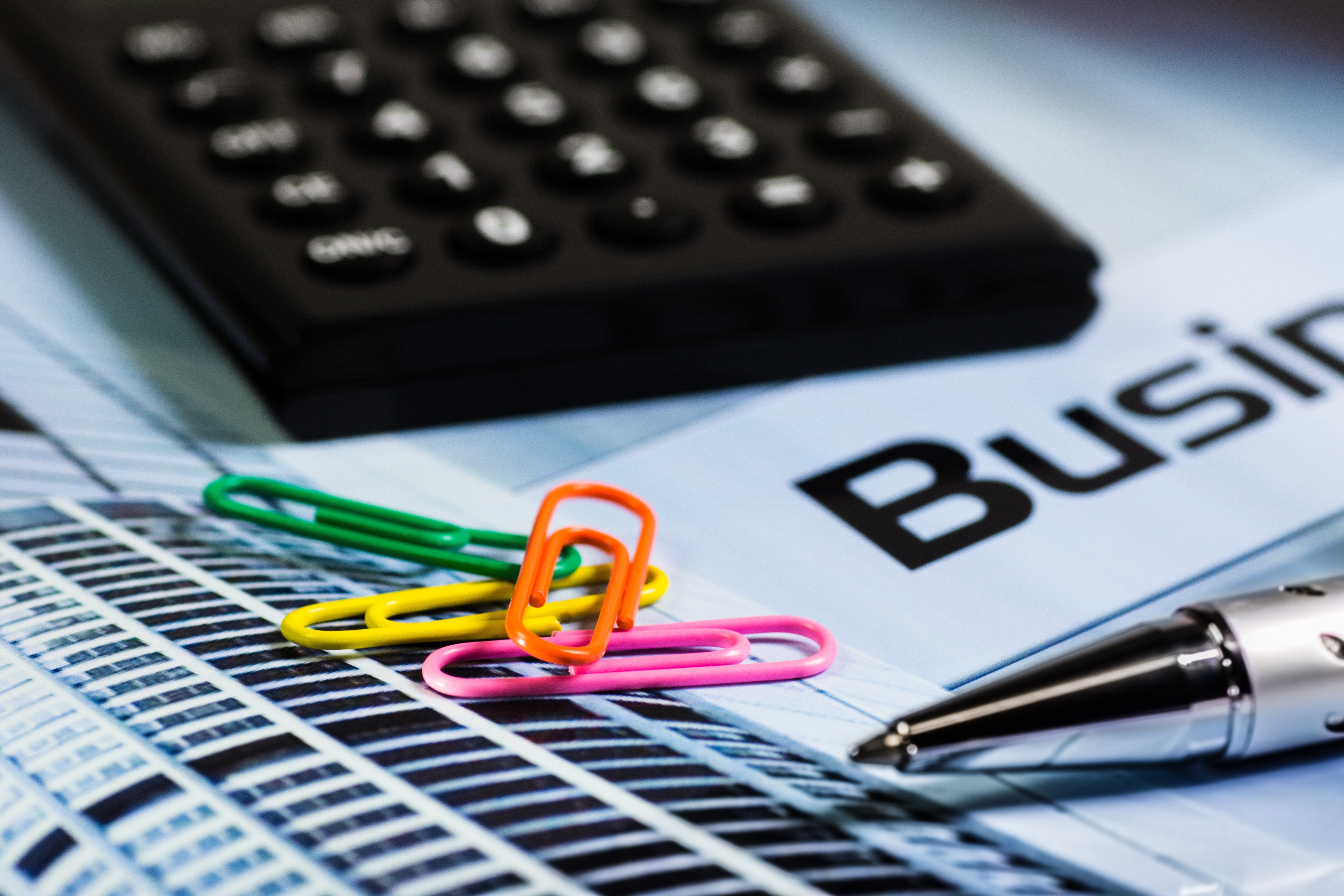 Black calculator sits above colored paper clips and a pen sitting on paper.