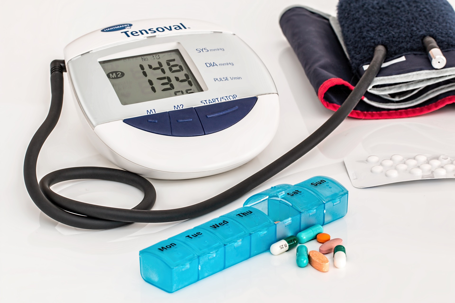White and blue blood pressure machine, open teal pill organizer and a blister pack of white tablets on a white surface.