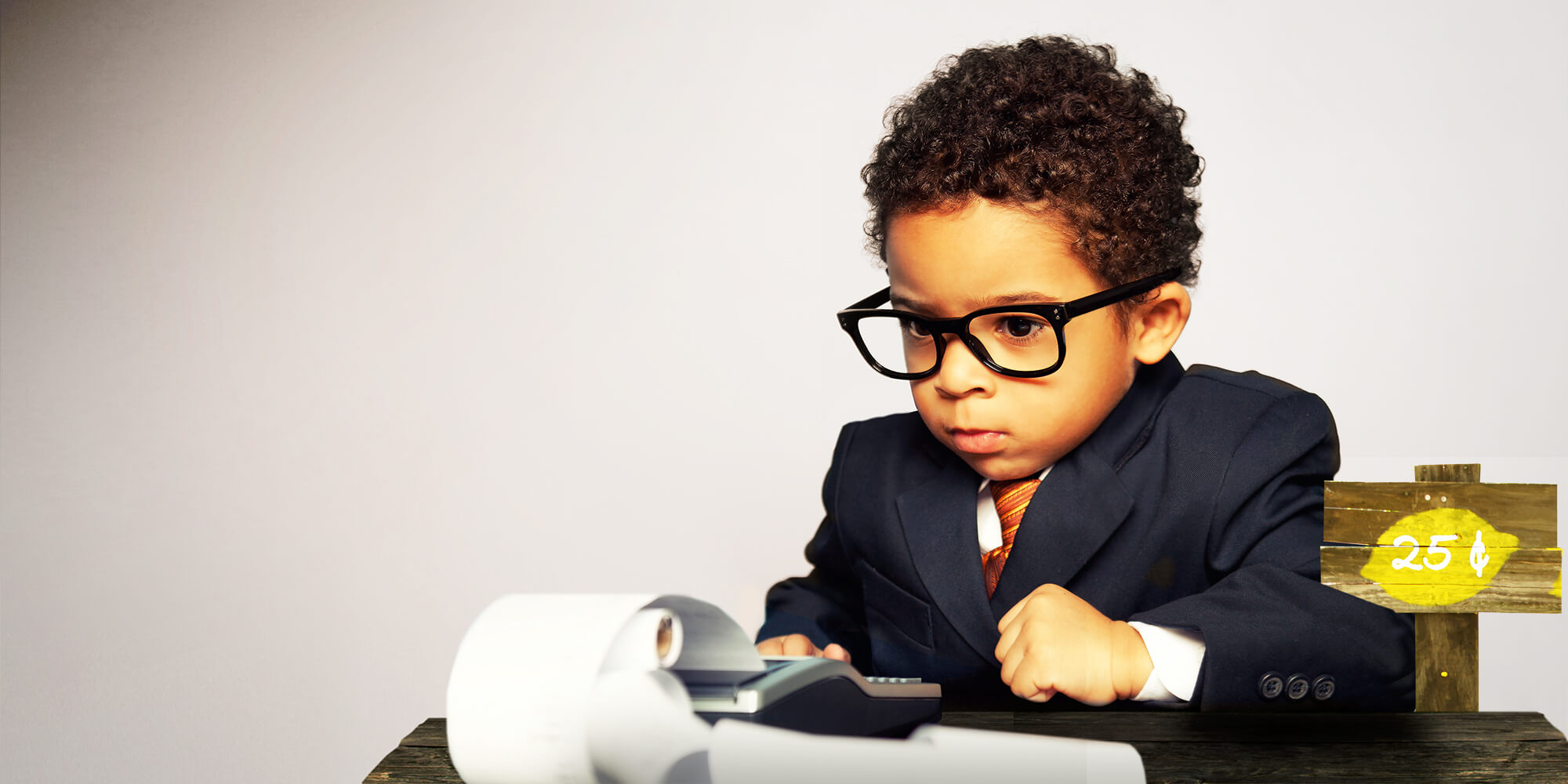 Curly haired boy dressed in a suit at a lemonade stand using a calculator.