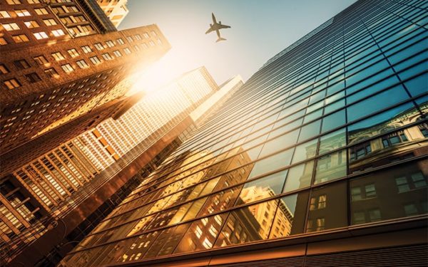 Reflections of city buildings in a large glass building with an airplane flies overhead.