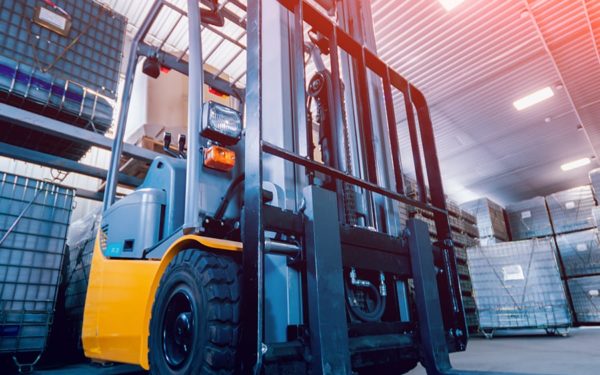 A gold and black fork lift sits in a warehouse in front of stacks of wrapped up pallets.