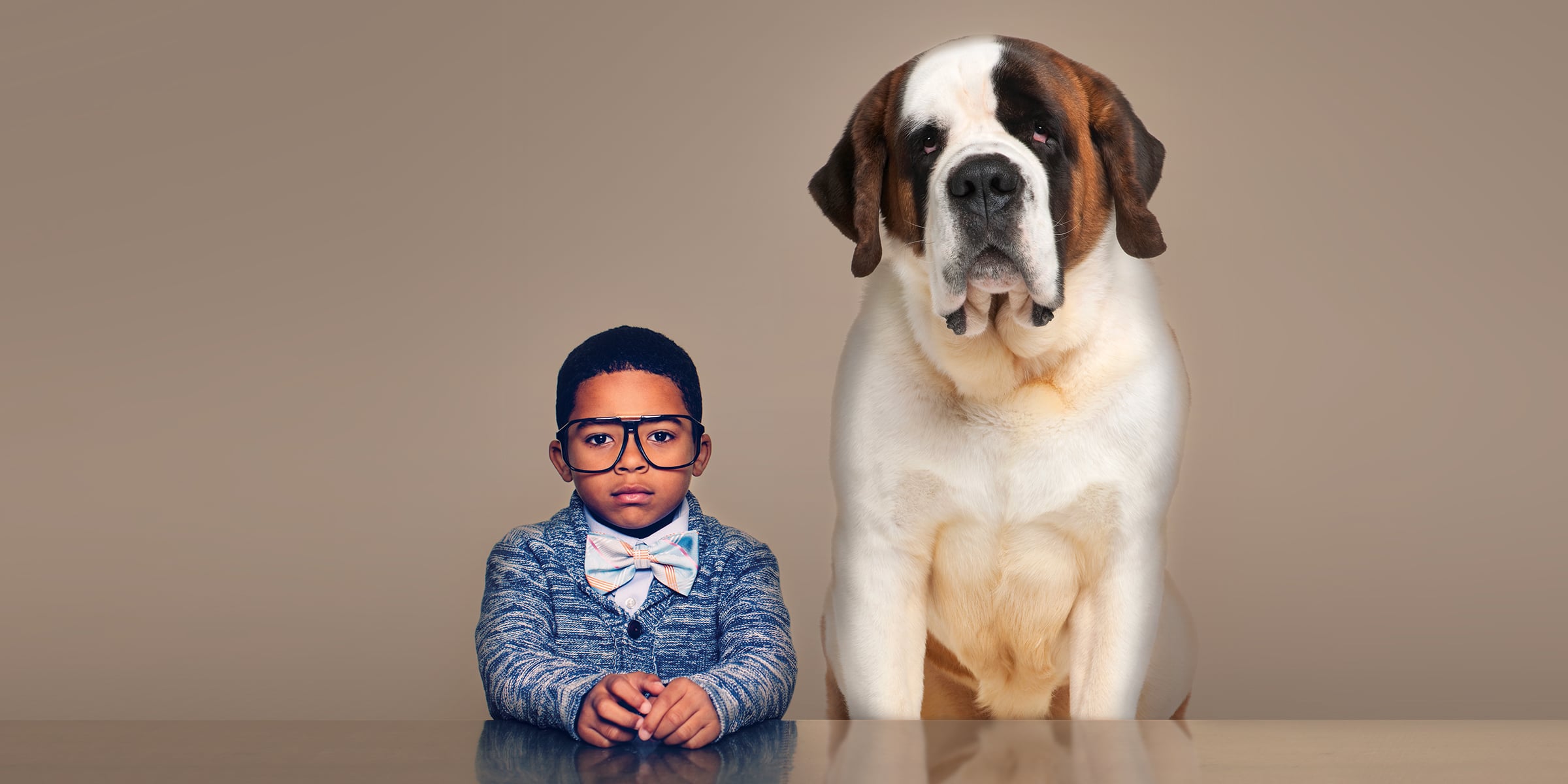 Child in cardigan, bow tie, and oversized glasses sits next to a St. Bernard at a table.