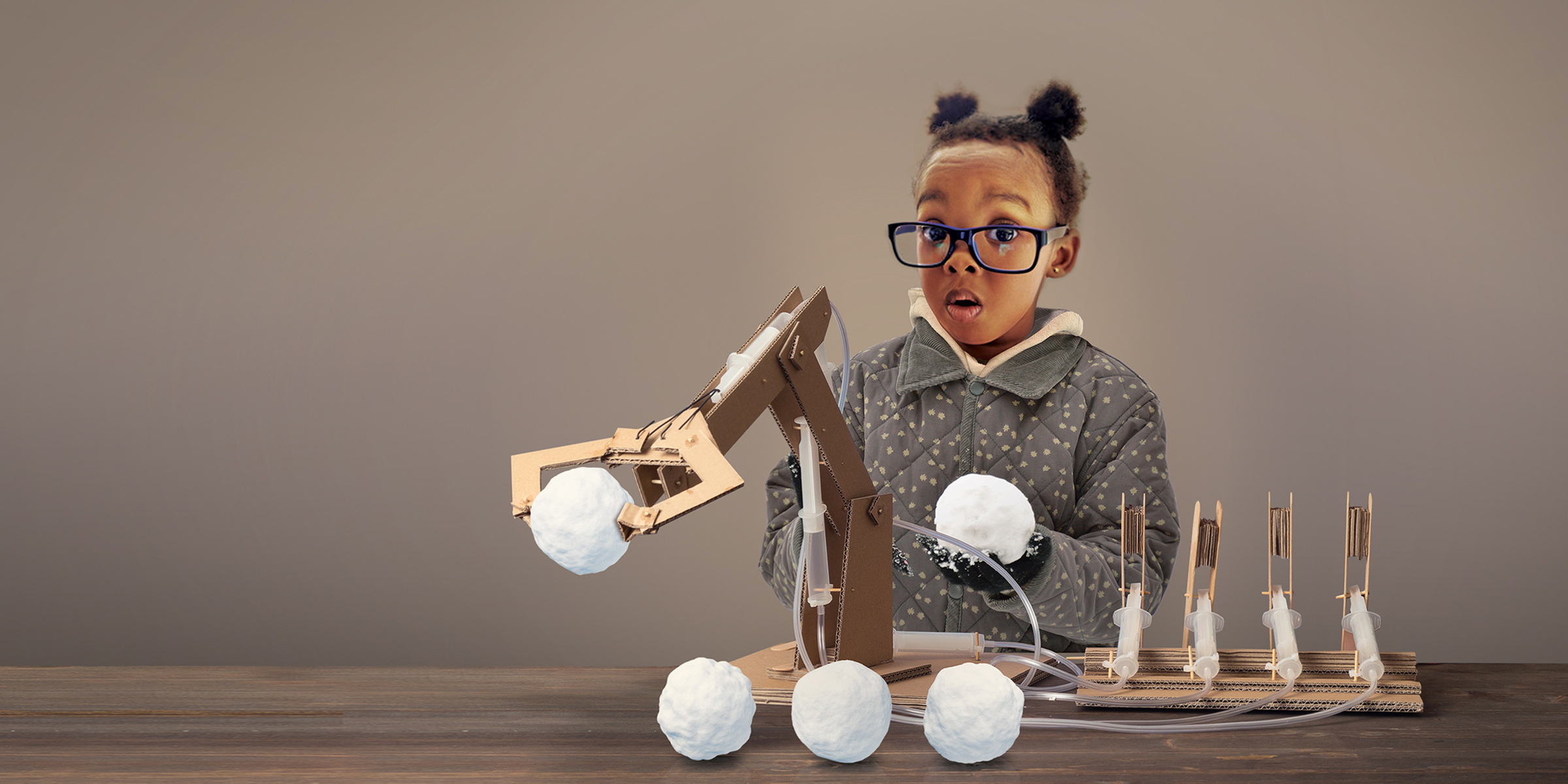 Little girl with oversized glasses holds a snow ball in her hand behind a homemade snowball making machine.