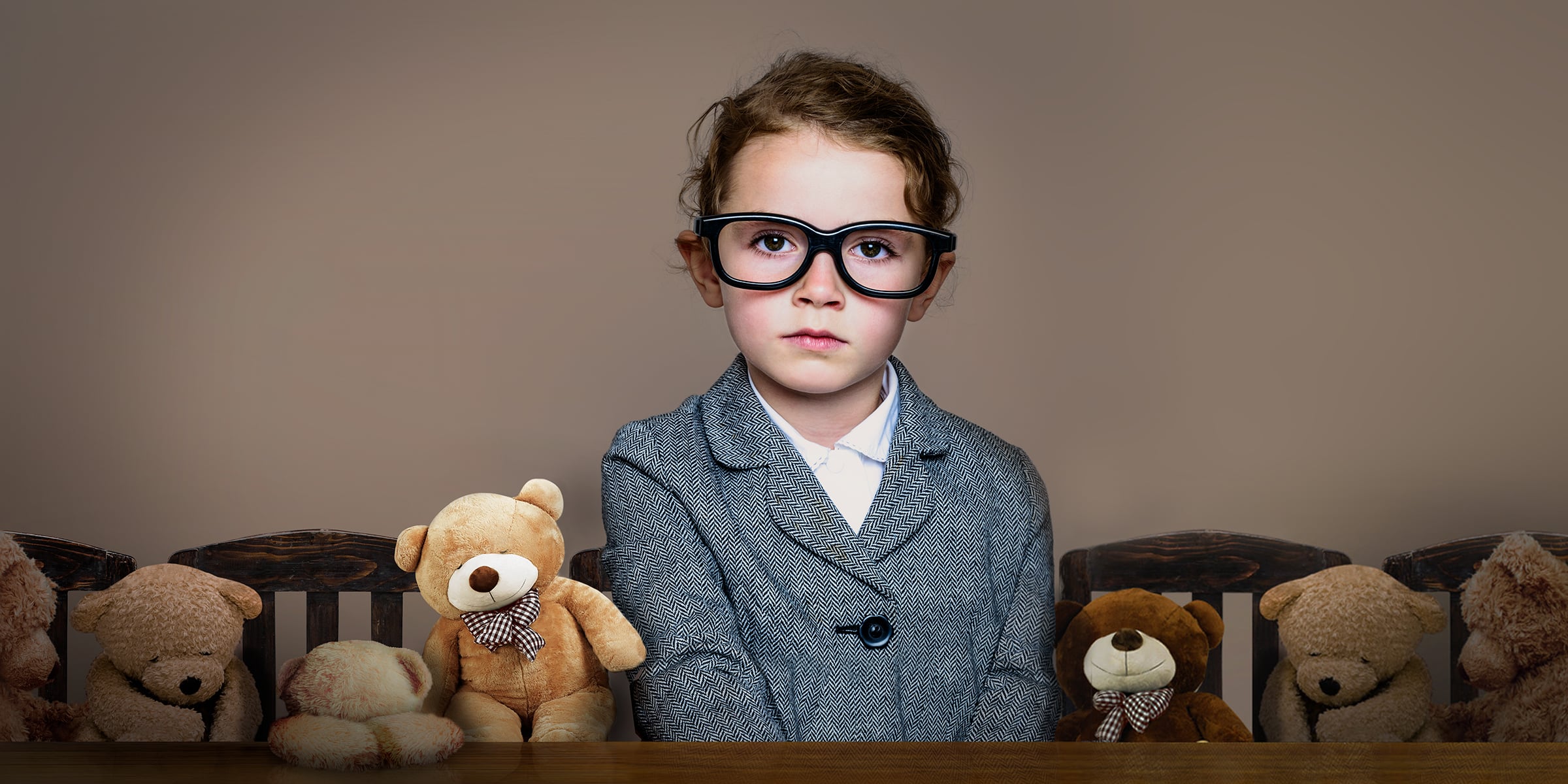 Child wearing a blazer and oversized glasses sits at a table, teddy bears sitting on either side of her.