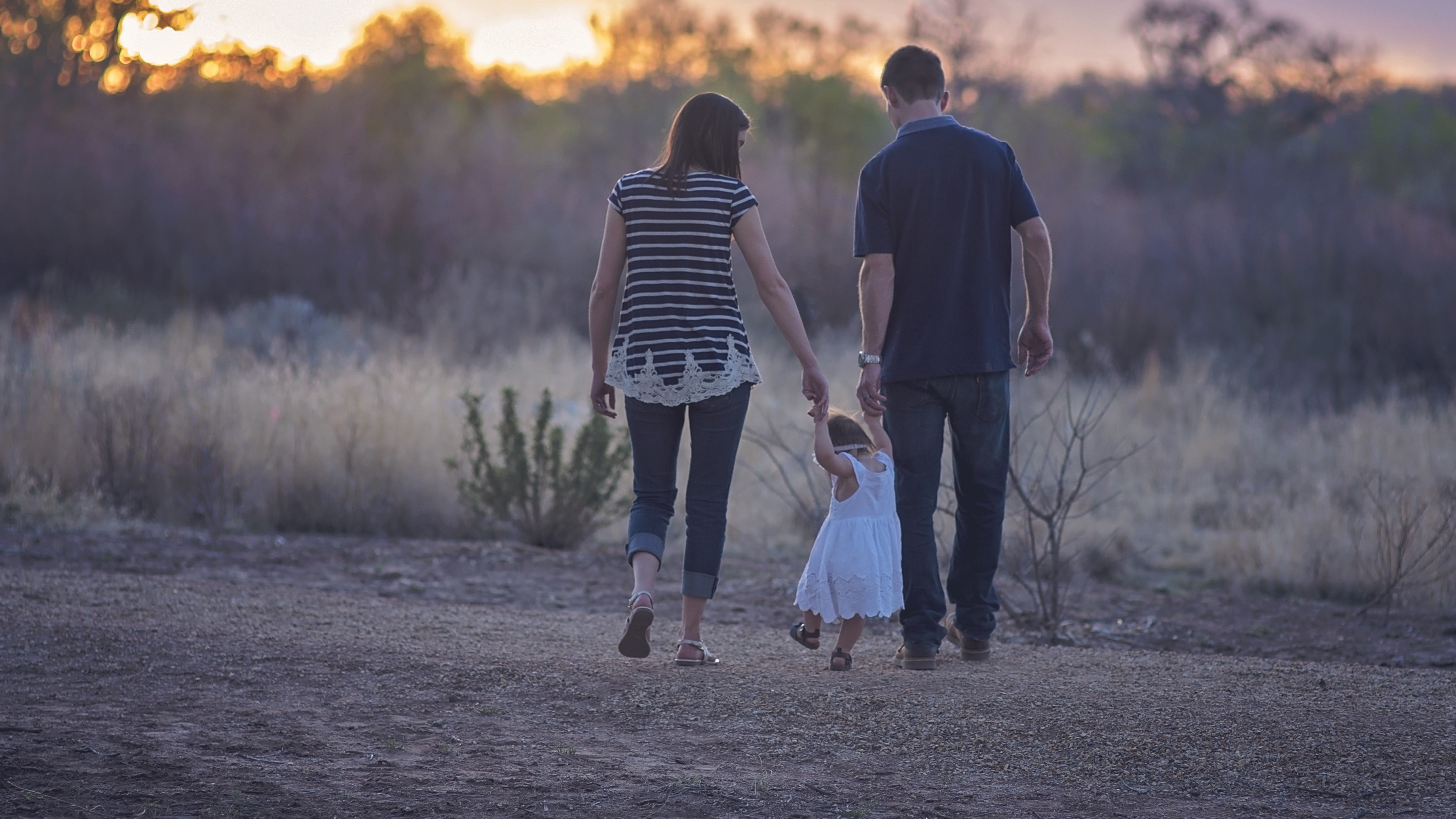 A man and a woman hold their toddlers hand while walking towards a sunset on a gravel path.