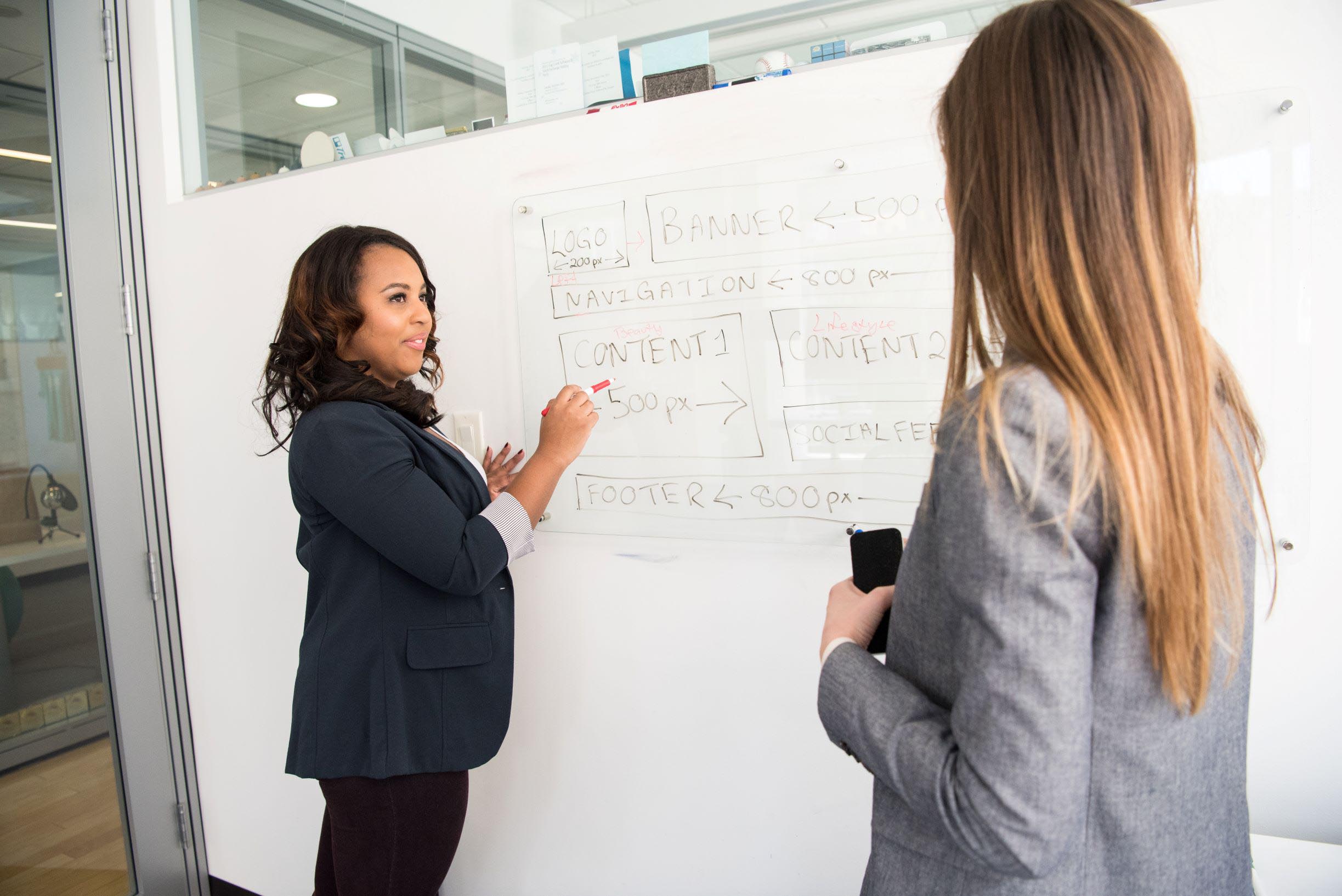 Two women stand in front of a white board with a flow chart on it.