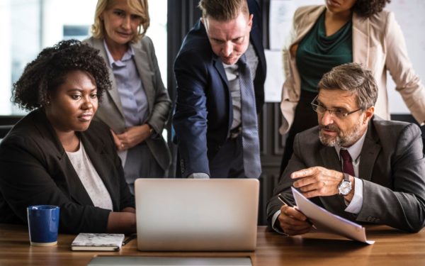 Five people dressed in business attire look at a laptop while in discussion.