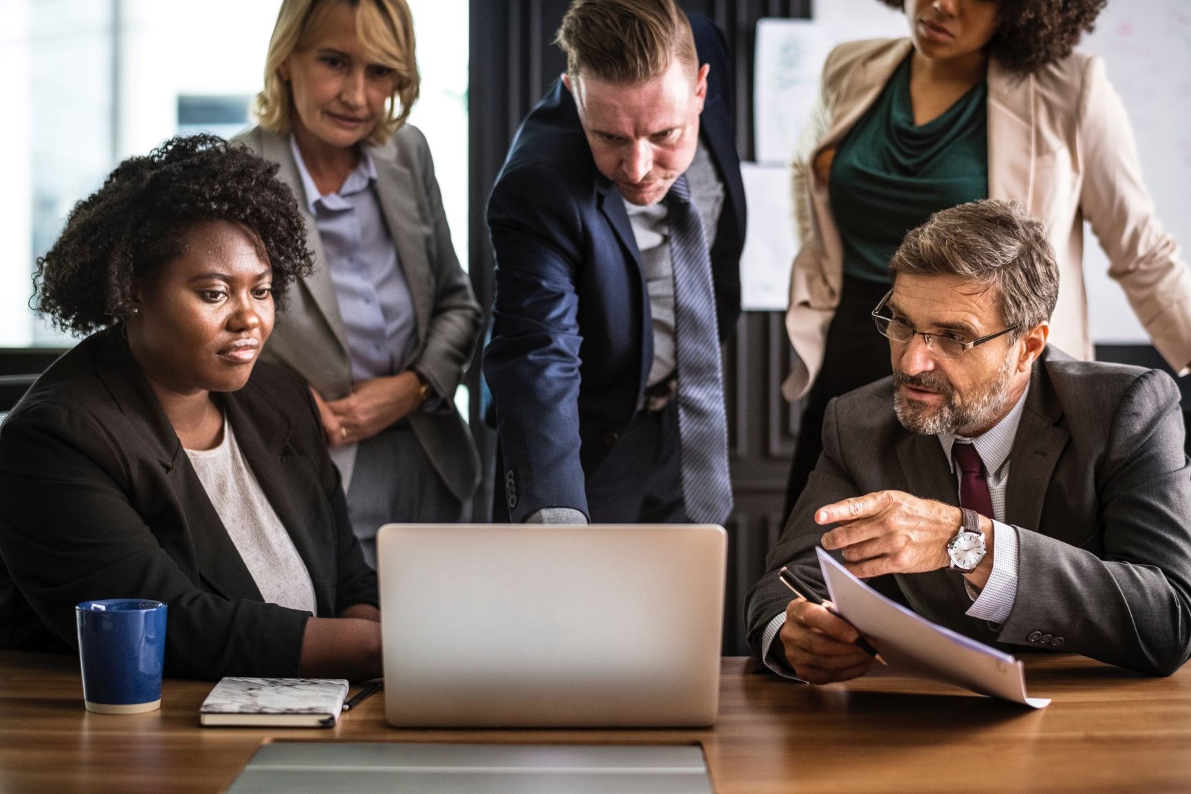 Five people dressed in business attire look at a laptop while in discussion.