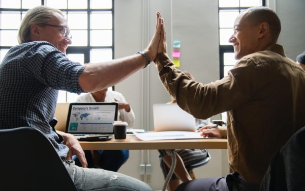 Two men smile while high fiving while sitting at a meeting.