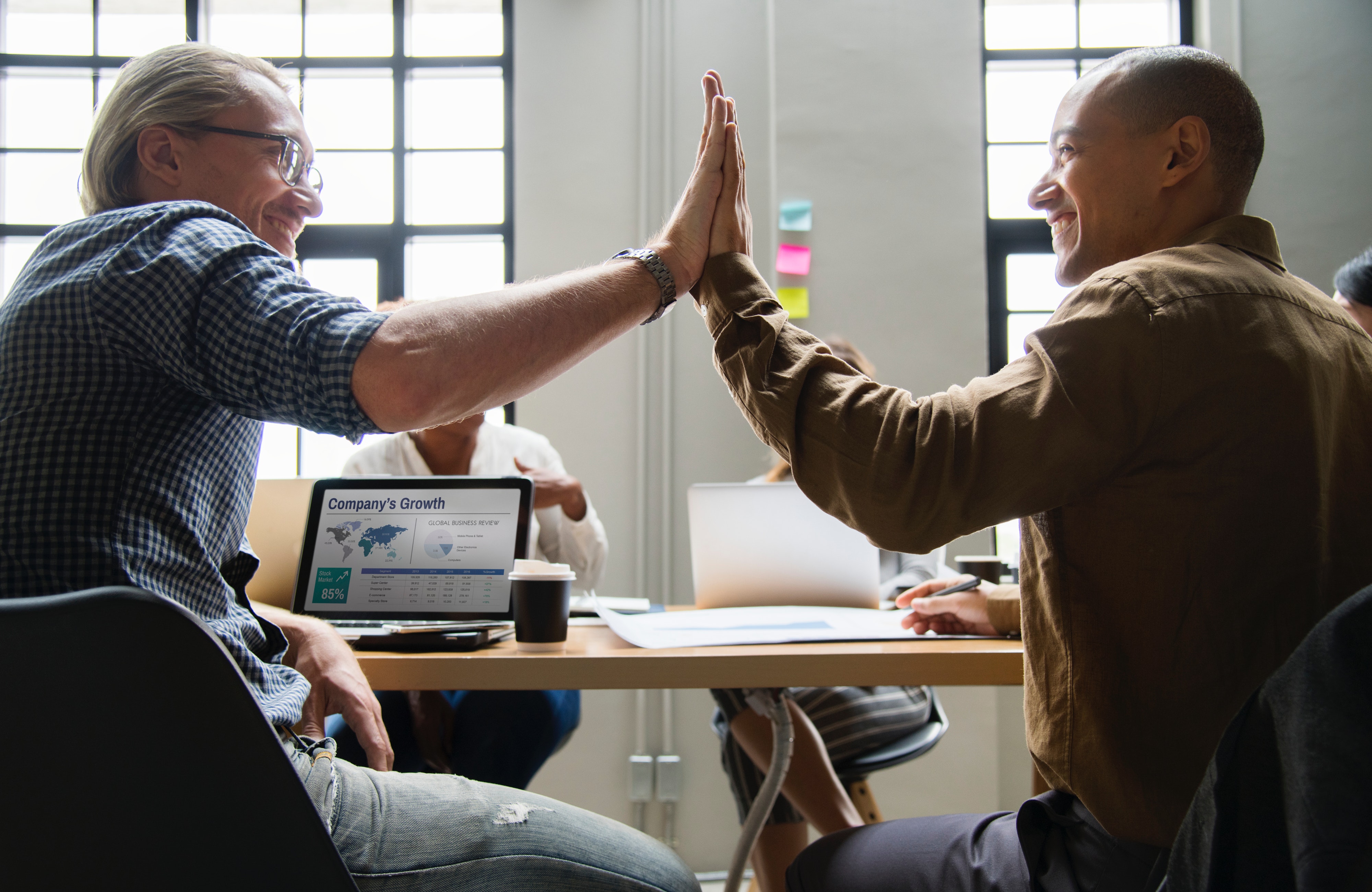 Two men smile while high fiving while sitting at a meeting.