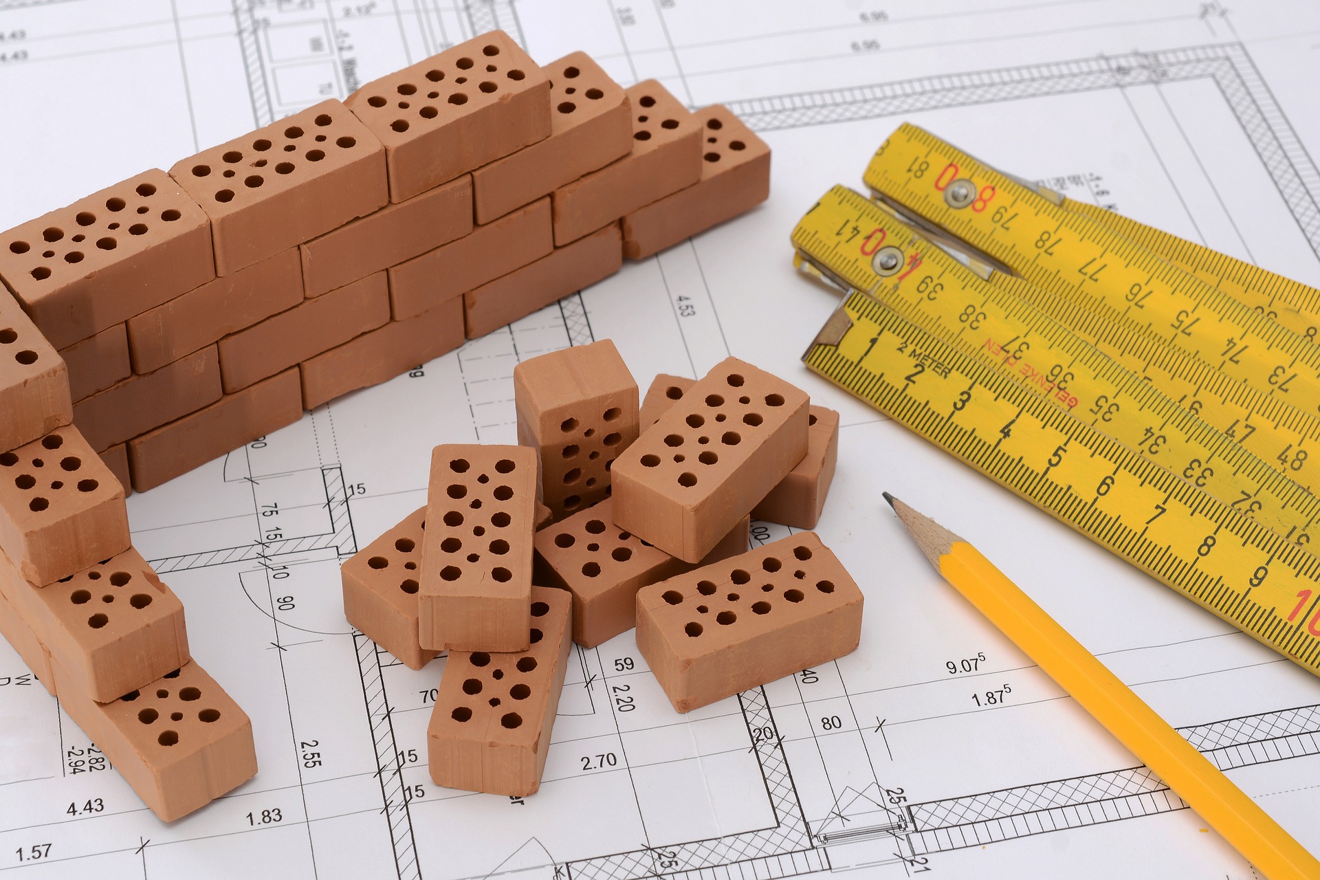 Miniature bricks form part of a structure next to a pencil and multiple tape measures on top of a blue print.