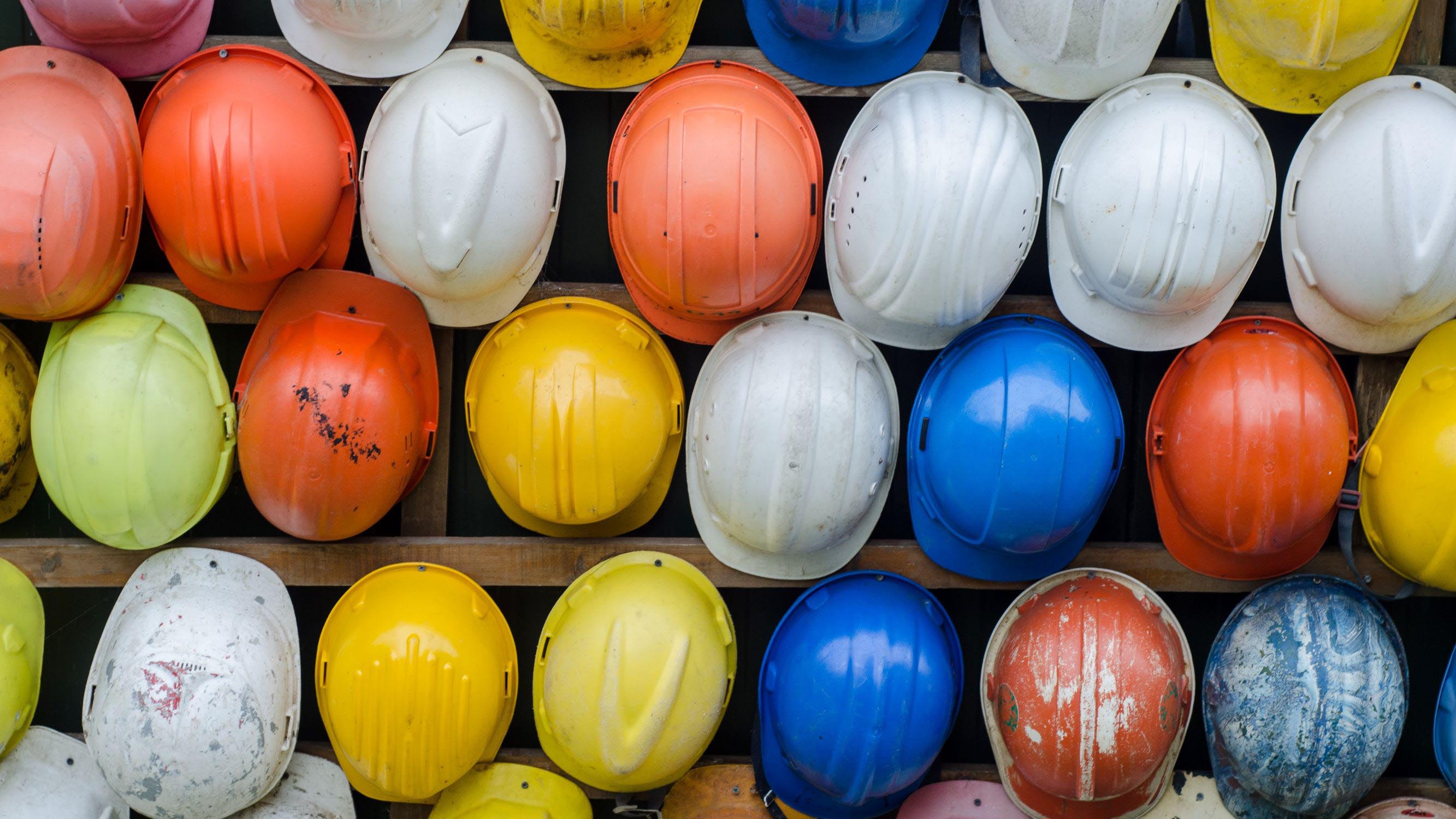 Blue, white, orange, and yellow hardhats hung up on a wooden structure.