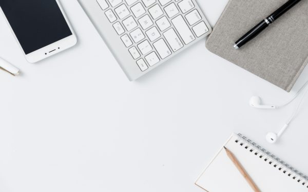 Flat lay of an iPhone, mac keyboard, pen, pencil, notebook, and ear buds on a white surface.