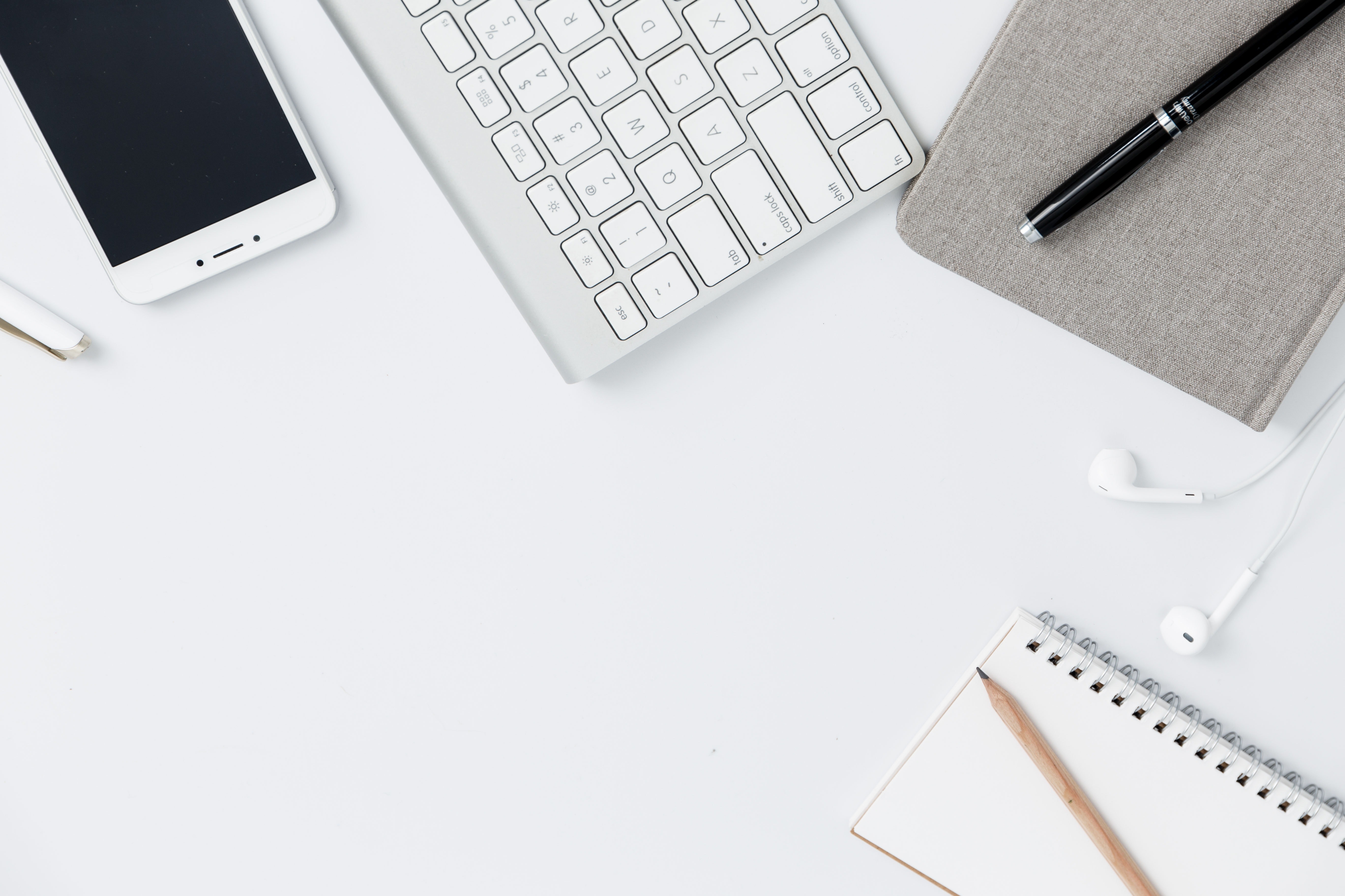 Flat lay of an iPhone, mac keyboard, pen, pencil, notebook, and ear buds on a white surface.