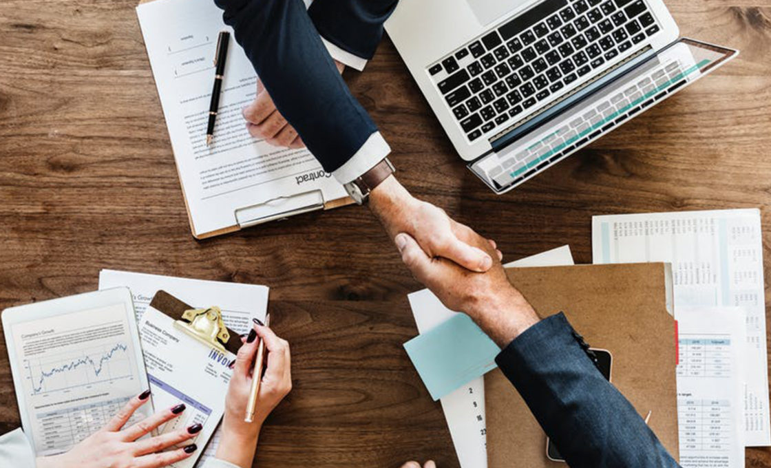 Two men in suits shake hands over top of a wooden table with papers, folders, and laptop on it.