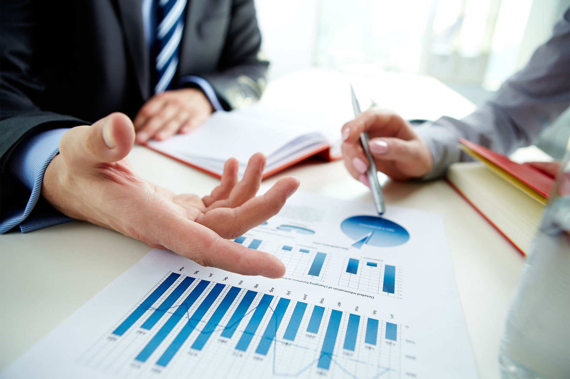 Man gestures to a document with various blue graphs sitting across from man holding a pen.