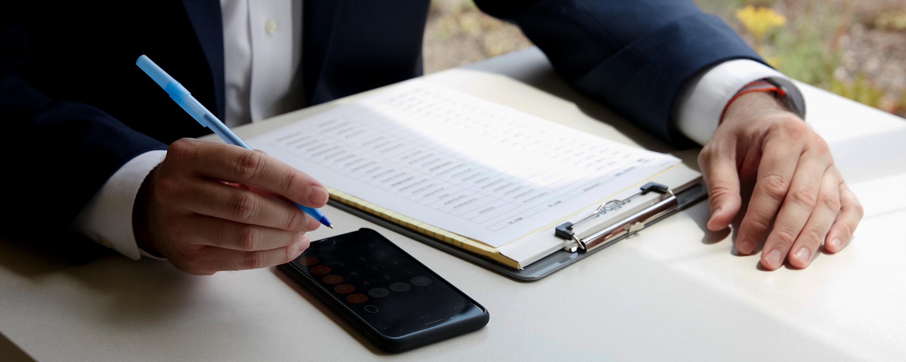 Man in a suit holds a pen above a table with a clipboard with papers on it and the calculator app open on his phone.