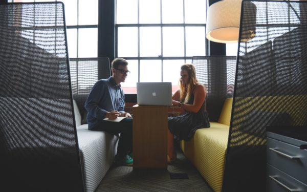 Man in glasses and woman in a dress sit in a work booth looking at laptop.