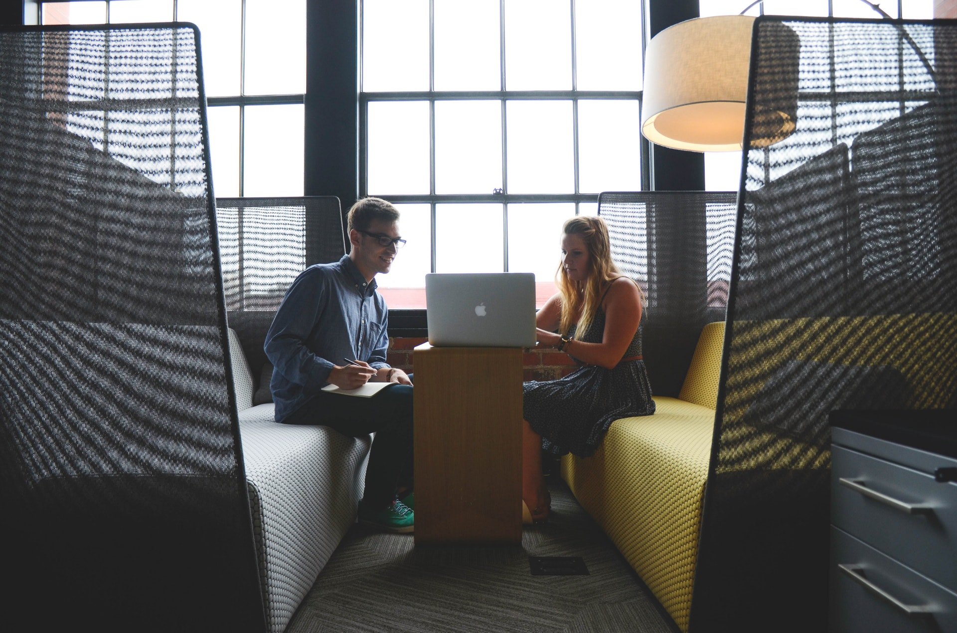 Man in glasses and woman in a dress sit in a work booth looking at laptop.