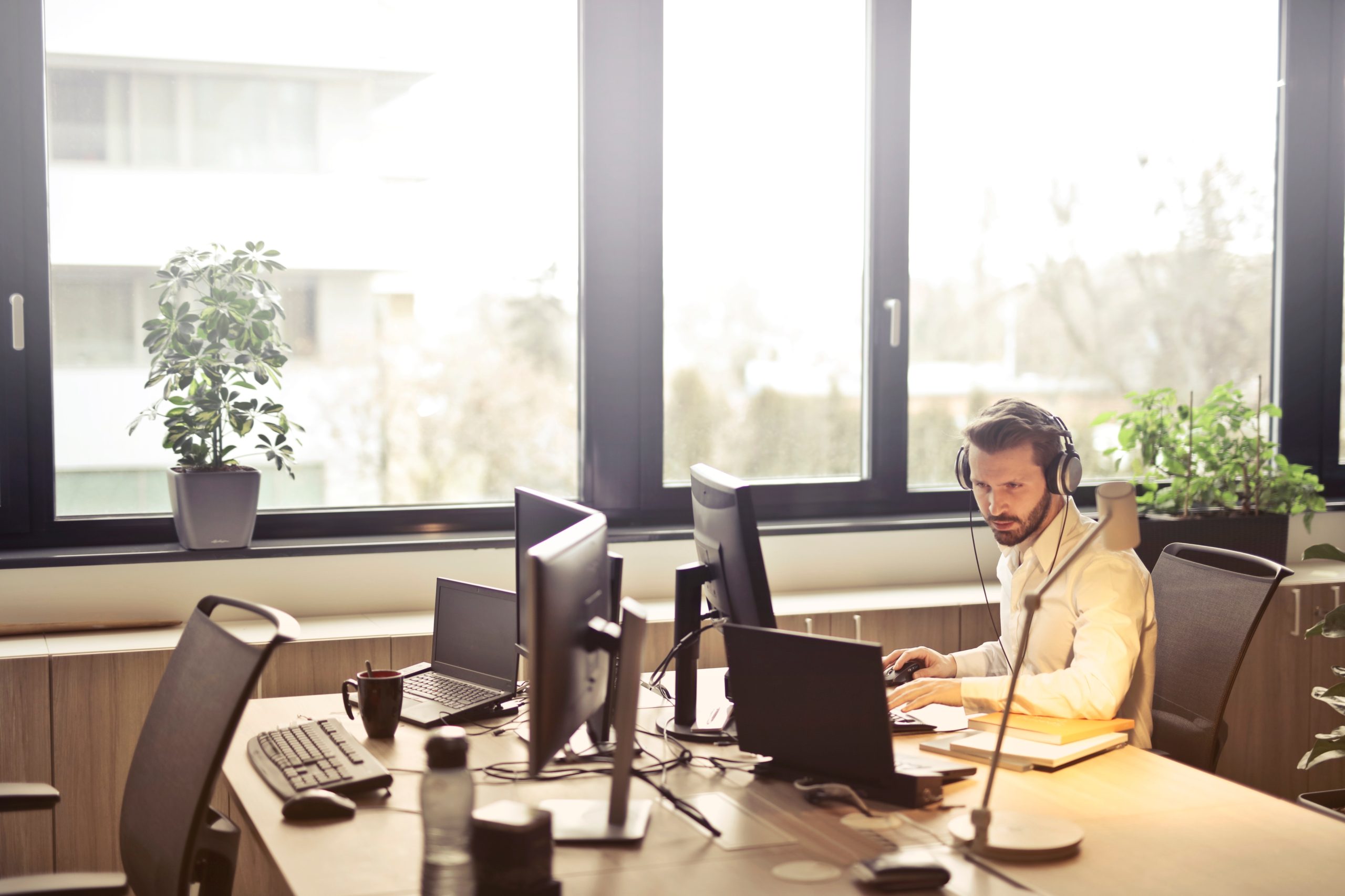 Man sits at desk on computer with headphones on in front of large glass windows.