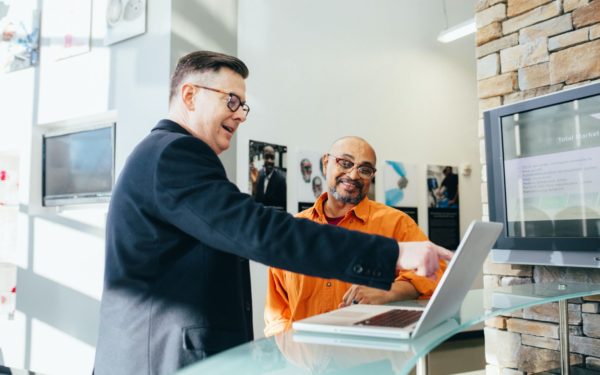 Man in orange shirt sits next to man in suit pointing at computer screen.