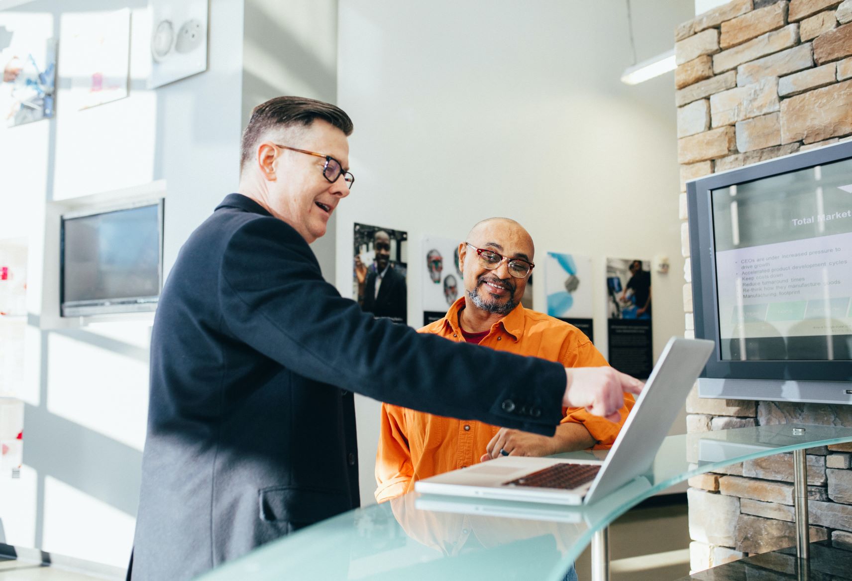 Man in orange shirt sits next to man in suit pointing at computer screen.