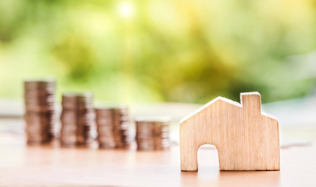 Miniature wooden cut out of a house with stacks of coins in the background.