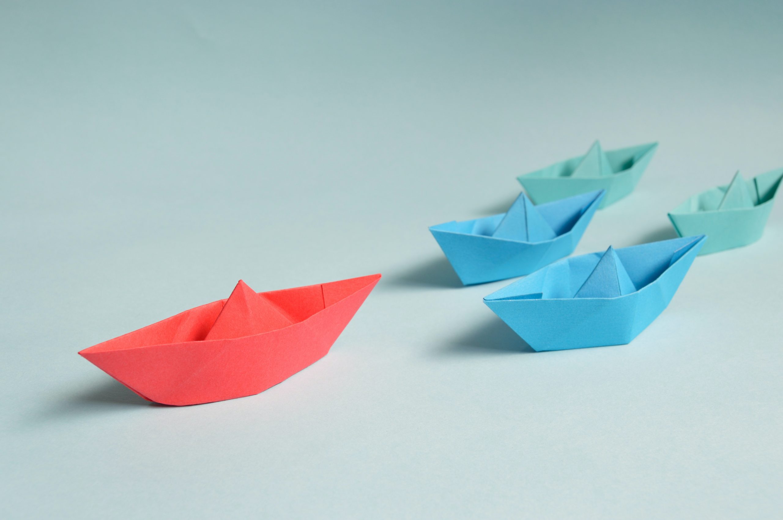 Red paper boat with two blue and two teal paper boats behind it on white surface.
