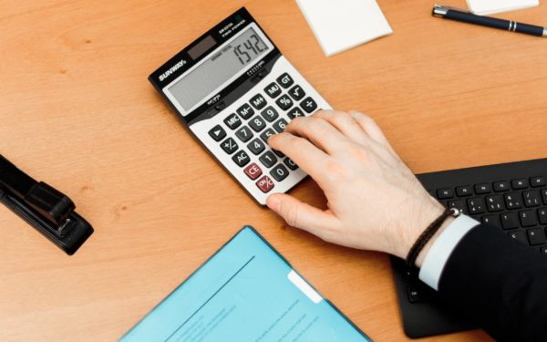 Two arms on a brown desk, one using a calculator while one arm rests on a blue folder.