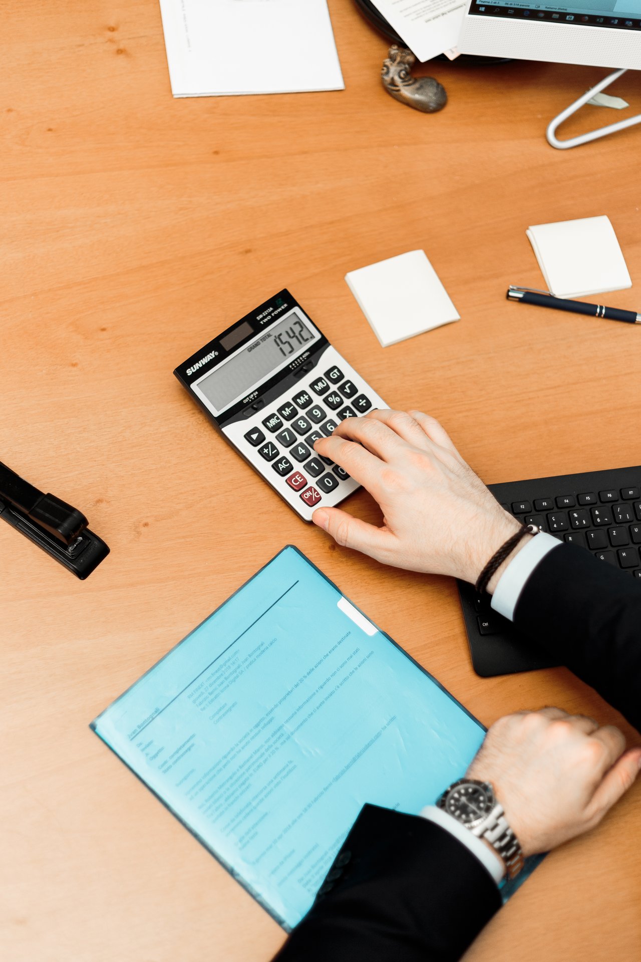 Two arms on a brown desk, one using a calculator while one arm rests on a blue folder.