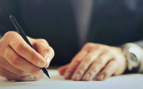 Hands of a man writing with pen sitting at desk.