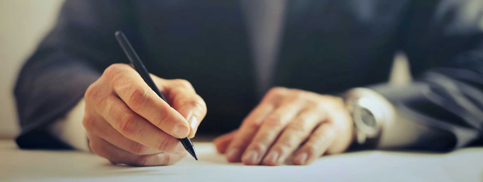 Hands of a man writing with pen sitting at desk.