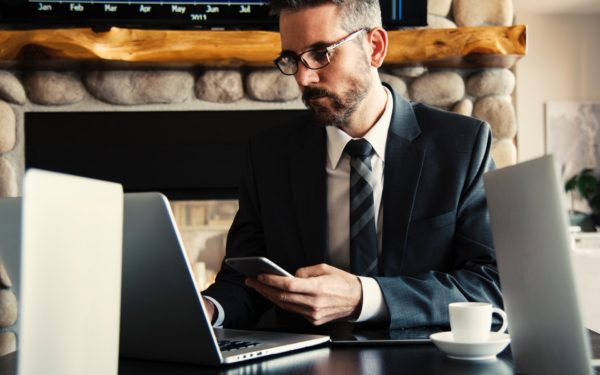 Man in glasses sitting in front of laptop holding cell phone.