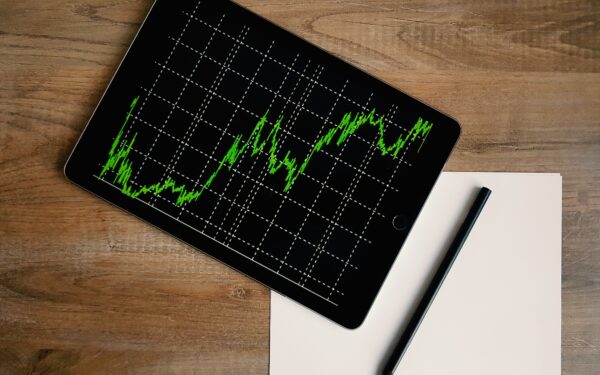 I pad with green line graph on screen sitting on a wooden desk on top of white paper.