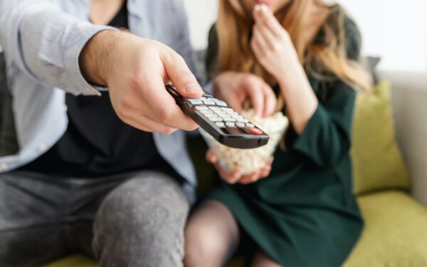 Man sitting on couch next to woman holding tv remote and bowl of popcorn.