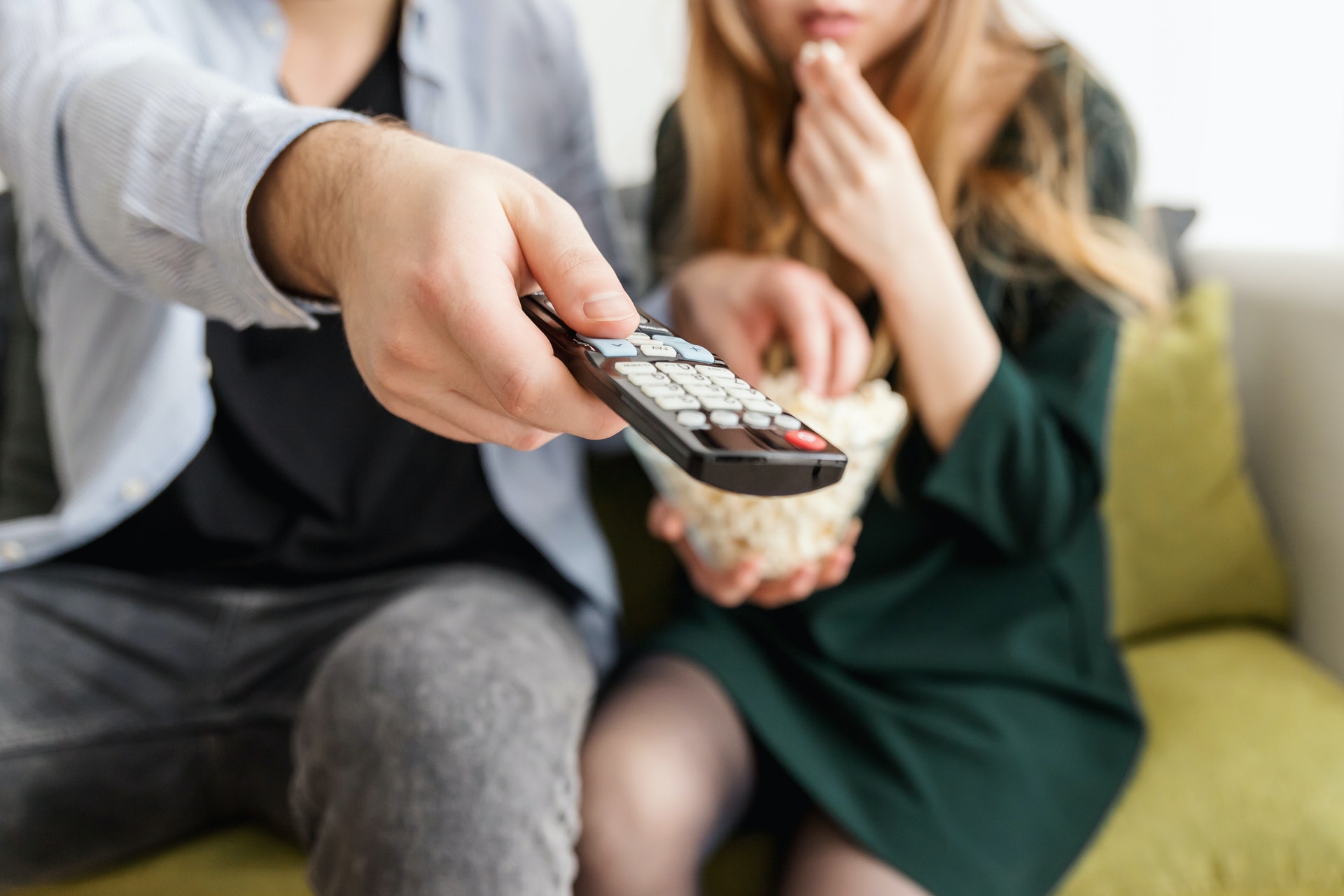 Man sitting on couch next to woman holding tv remote and bowl of popcorn.