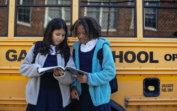 Two girls dressed in school uniforms reading a textbook next to a school bus.