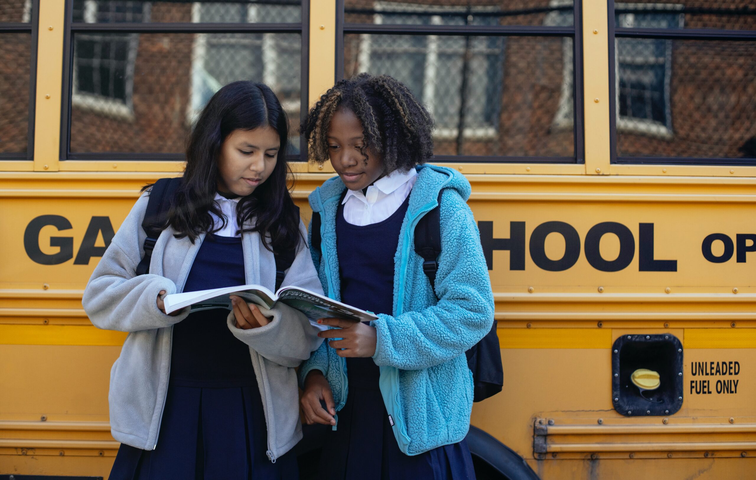 Two girls dressed in school uniforms reading a textbook next to a school bus.