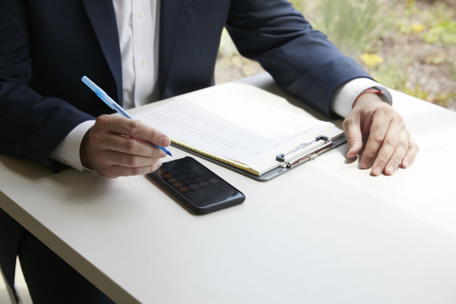 Man sitting at table with calculator app on phone and clipboard.