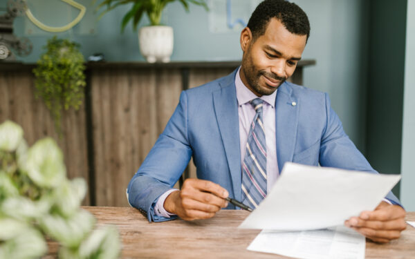 A man sits in a light blue suit holding a paper and pen sitting at a wooden desk.