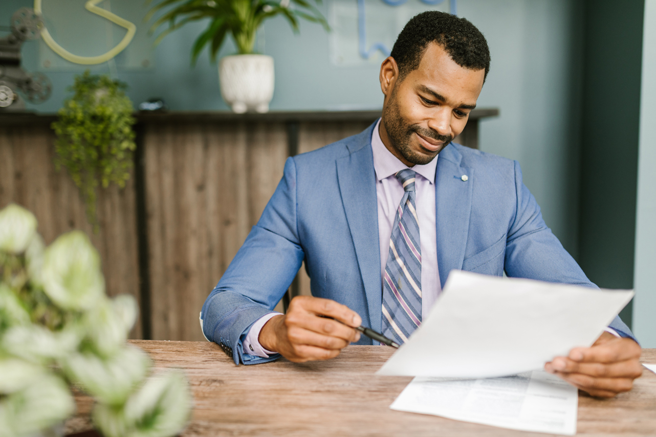 A man sits in a light blue suit holding a paper and pen sitting at a wooden desk.