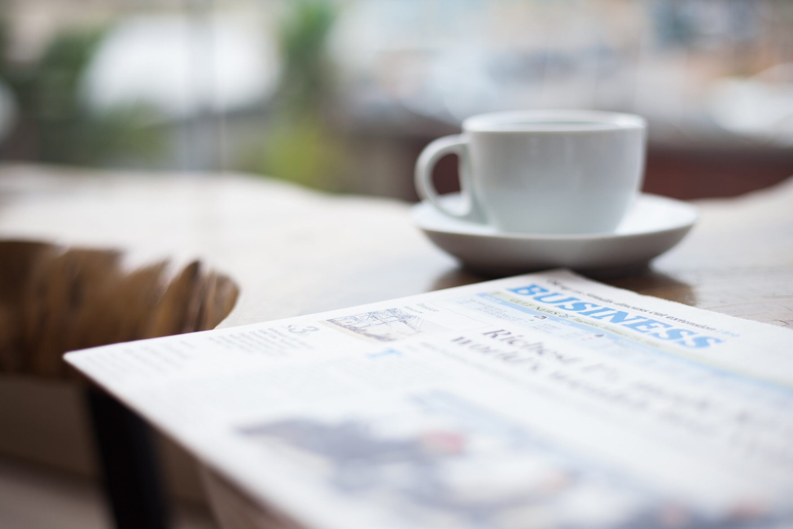 Business section of newspaper sitting on desk next to white cup and saucer.