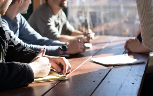 Three people sitting at table with pens in hand and one person standing up on opposite side of table.