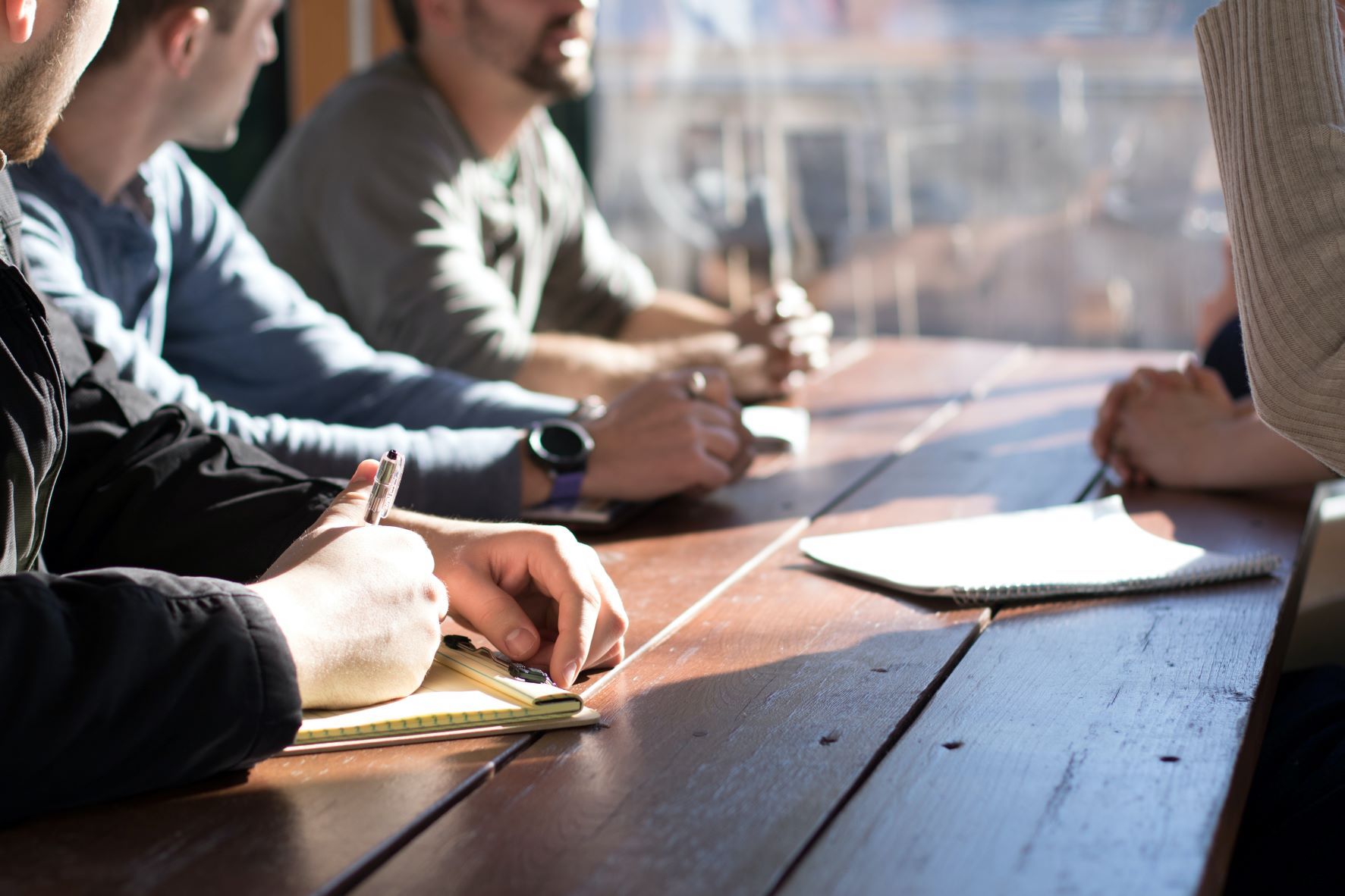 Three people sitting at table with pens in hand and one person standing up on opposite side of table.