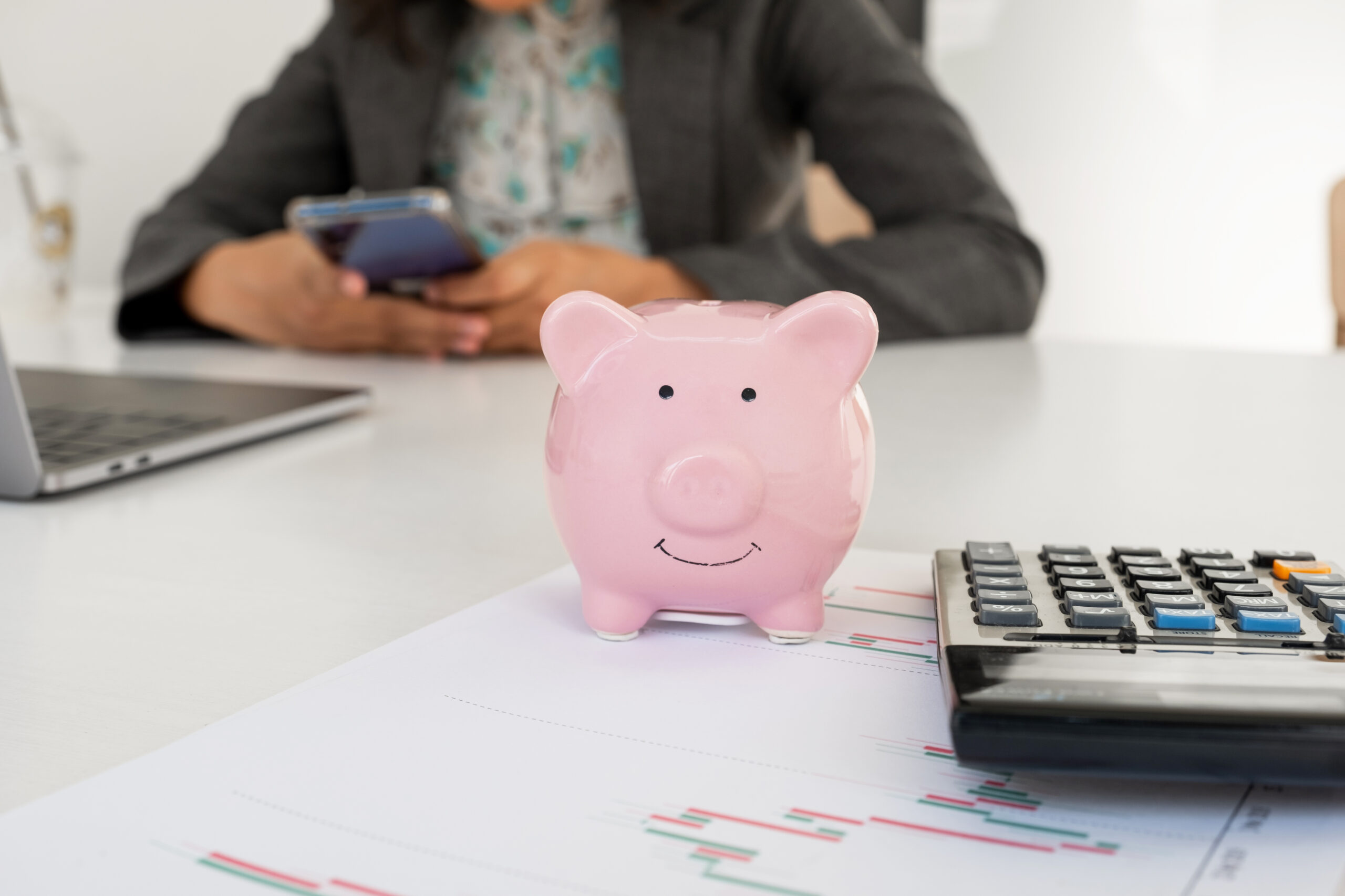 Person holding cell phone in their hands at desk with pink piggy bank calculator and papers.