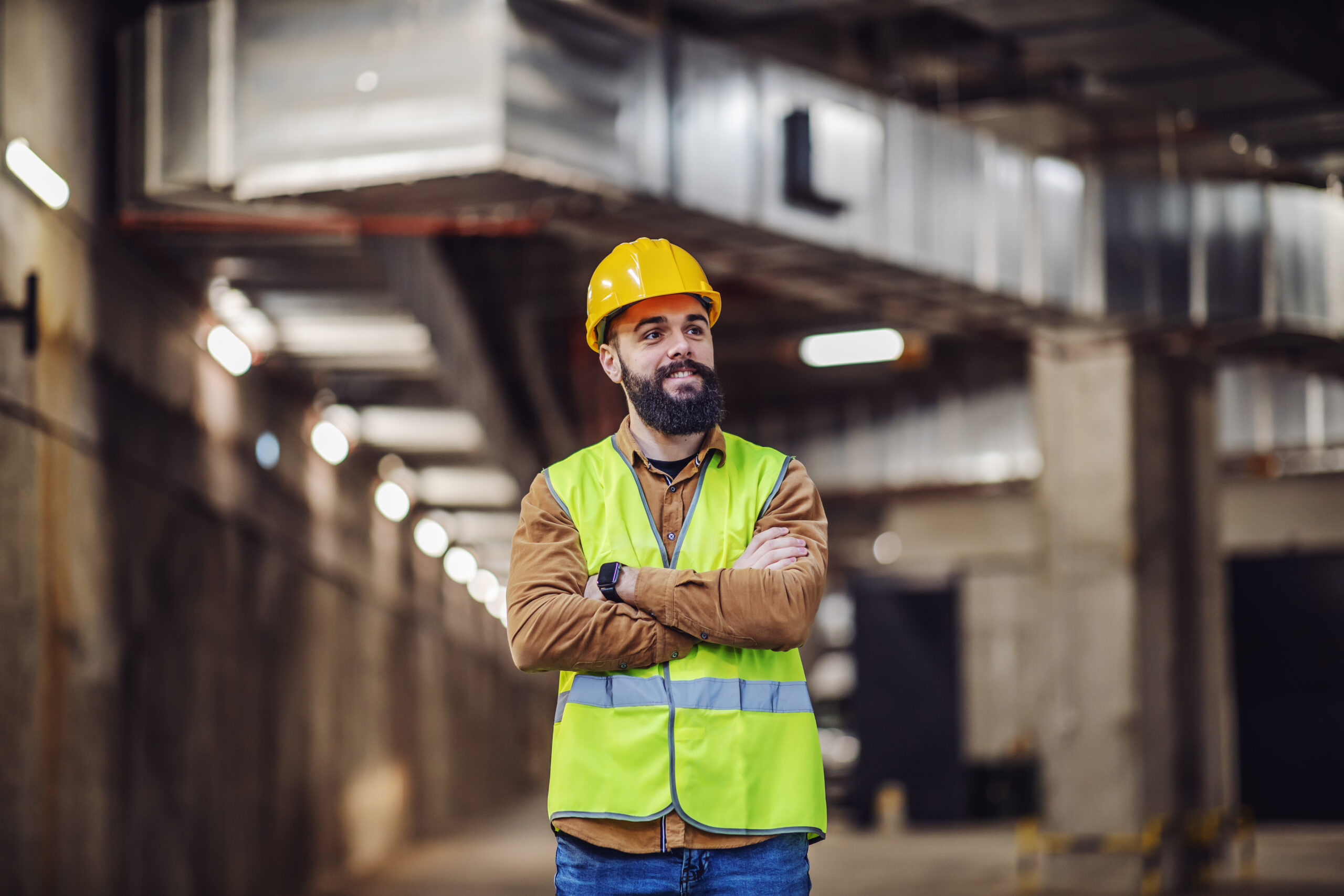 Young proud smiling attractive bearded supervisor standing in building in construction process with arms folded and overseeing his workers.