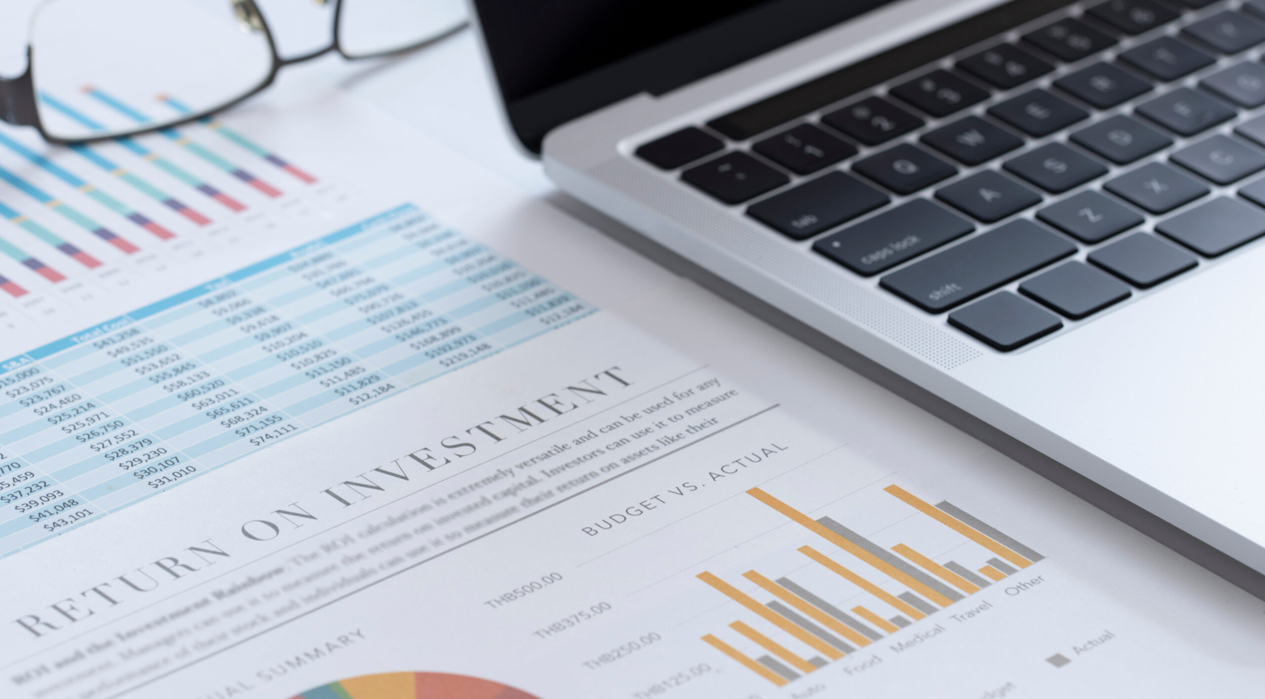 Paper with numbers and charts next to an open laptop and eyeglasses.