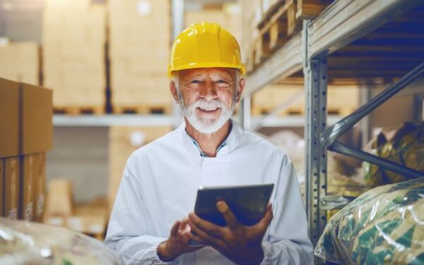 Smiling man in yellow helmet holding tablet in a warehouse.
